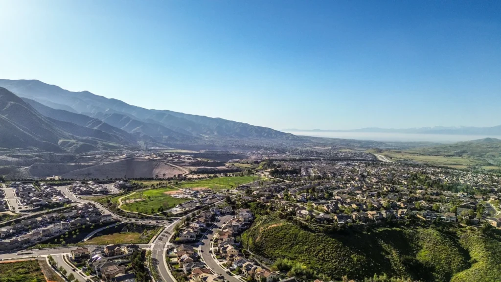 Aerial view of a sprawling suburban town in a valley with green hills in the foreground and mountains in the distance under a clear blue sky