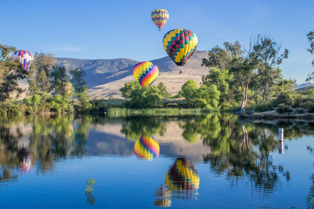 Several colorfully patterned hot air balloons hover above a calm lake with green trees and distant hills under a clear blue sky, reflecting in the water.